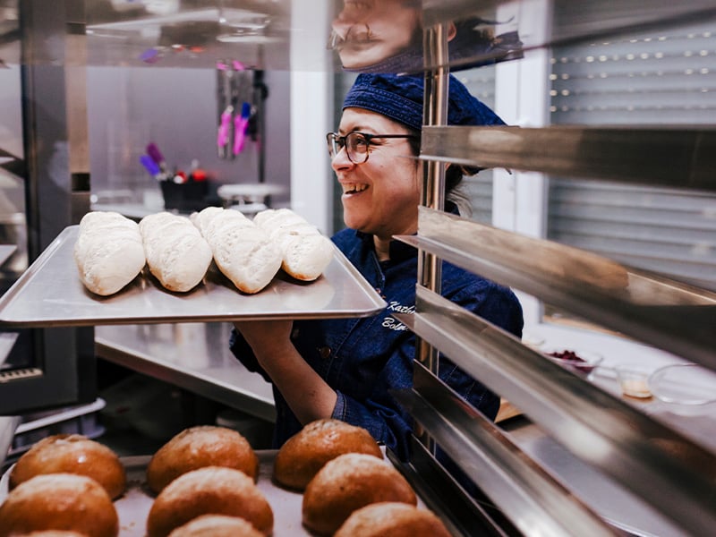 Smiling baker places a tray of fresh bread onto bakery racks, taking pride in her craft