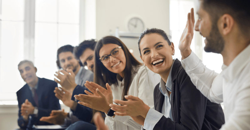 A diverse group of employees, smiling and cheering, celebrate a coworker's achievement.
