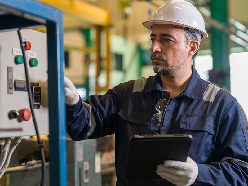 Attentive technician in coveralls inspects a control panel while holding a tablet