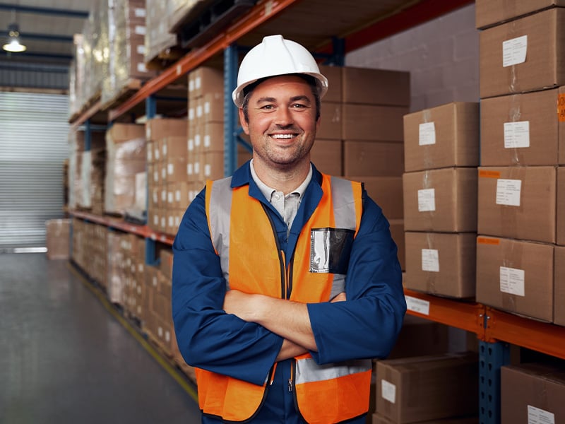 Confident warehouse supervisor stands arms crossed among stocked shelves, embodying reliable leadership