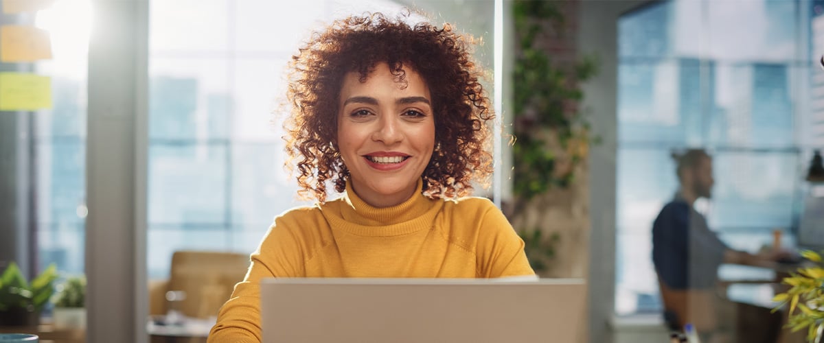 Happy employee smiling at her laptop in a bright, plant-filled modern office