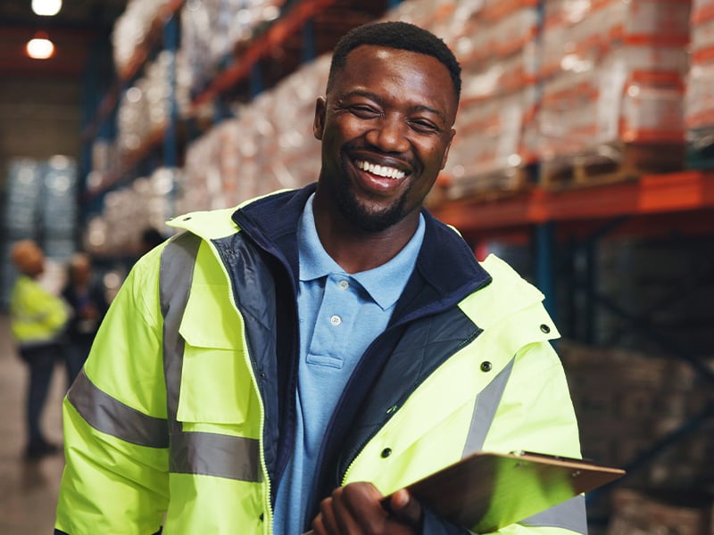Joyful warehouse worker in safety vest holding a clipboard on the floor