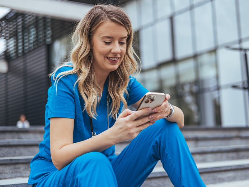 Nurse in scrubs smiling while checking her phone on hospital steps outside
