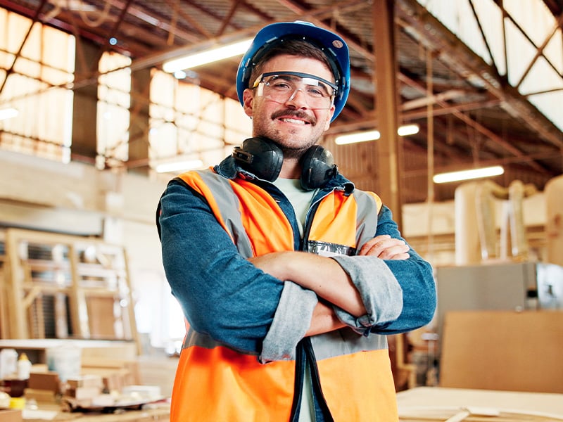 Proud manufacturing worker in hard hat and safety gear smiling arms crossed on the floor