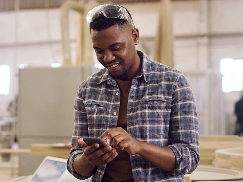 Smiling employee in a workshop checks his phone, blending craftsmanship with everyday connection
