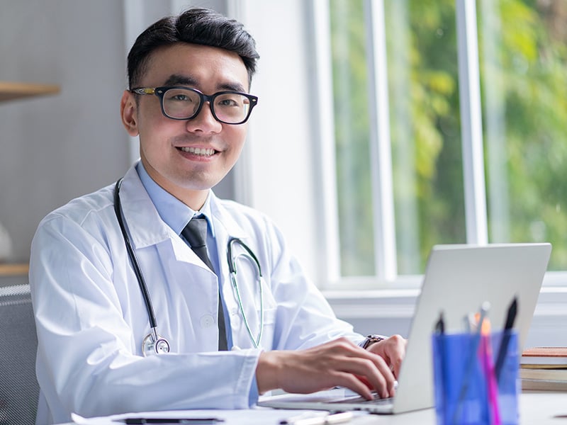Smiling physician at his desk works on a laptop, dedicated to quality patient care