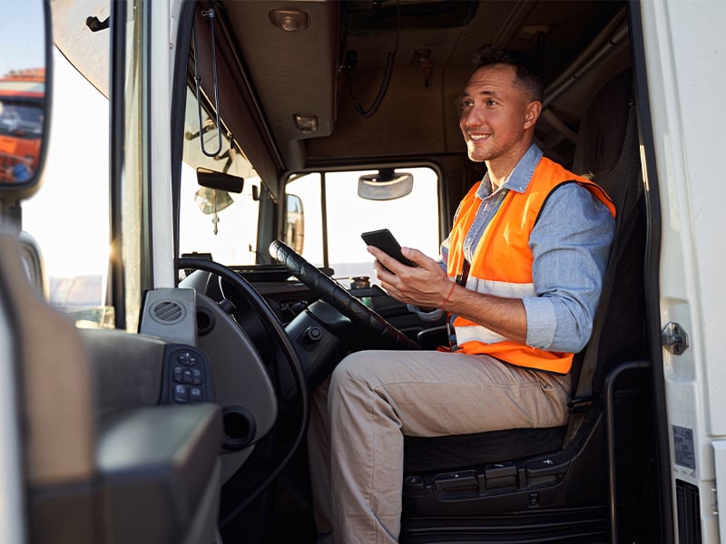 Smiling truck driver in safety vest checking his phone in the cab