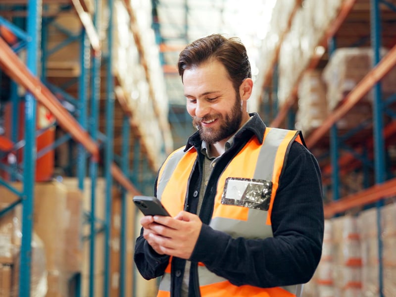 Smiling warehouse worker in safety vest checking his phone on the floor