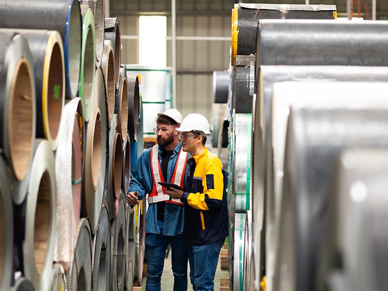 Two safety-vested workers review a tablet while inspecting stacked steel coils