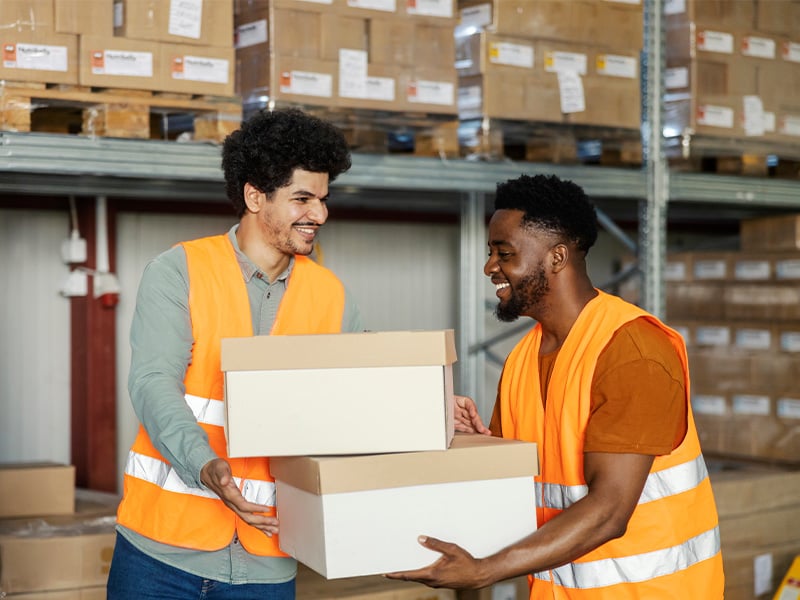 Two warehouse teammates in safety vests smiling while working together