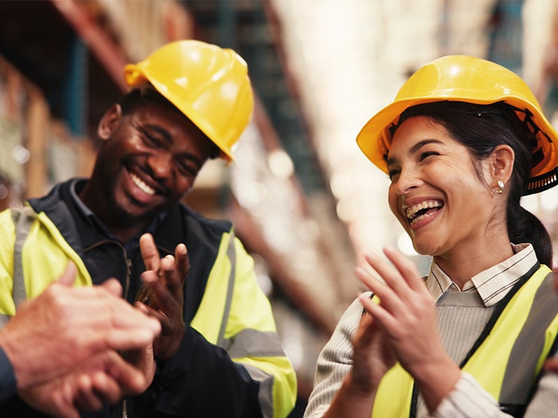 Warehouse team in hard hats laughing and clapping together in a celebratory moment