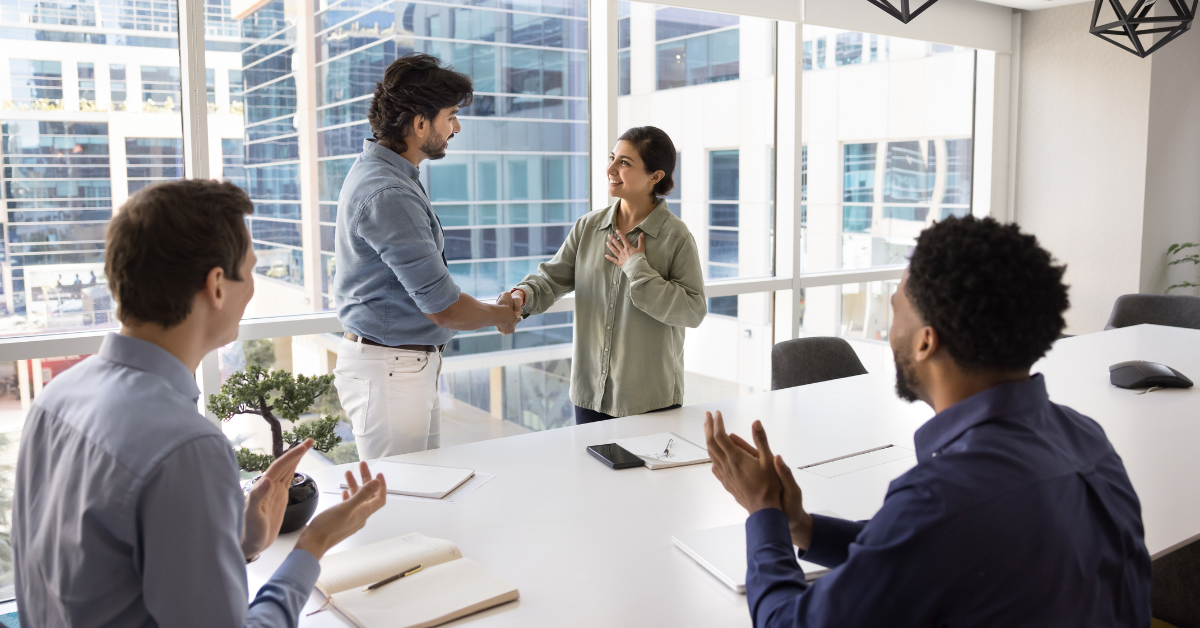 A woman and a man shake hands at the front of a conference room while two colleagues seated at the table applaud.